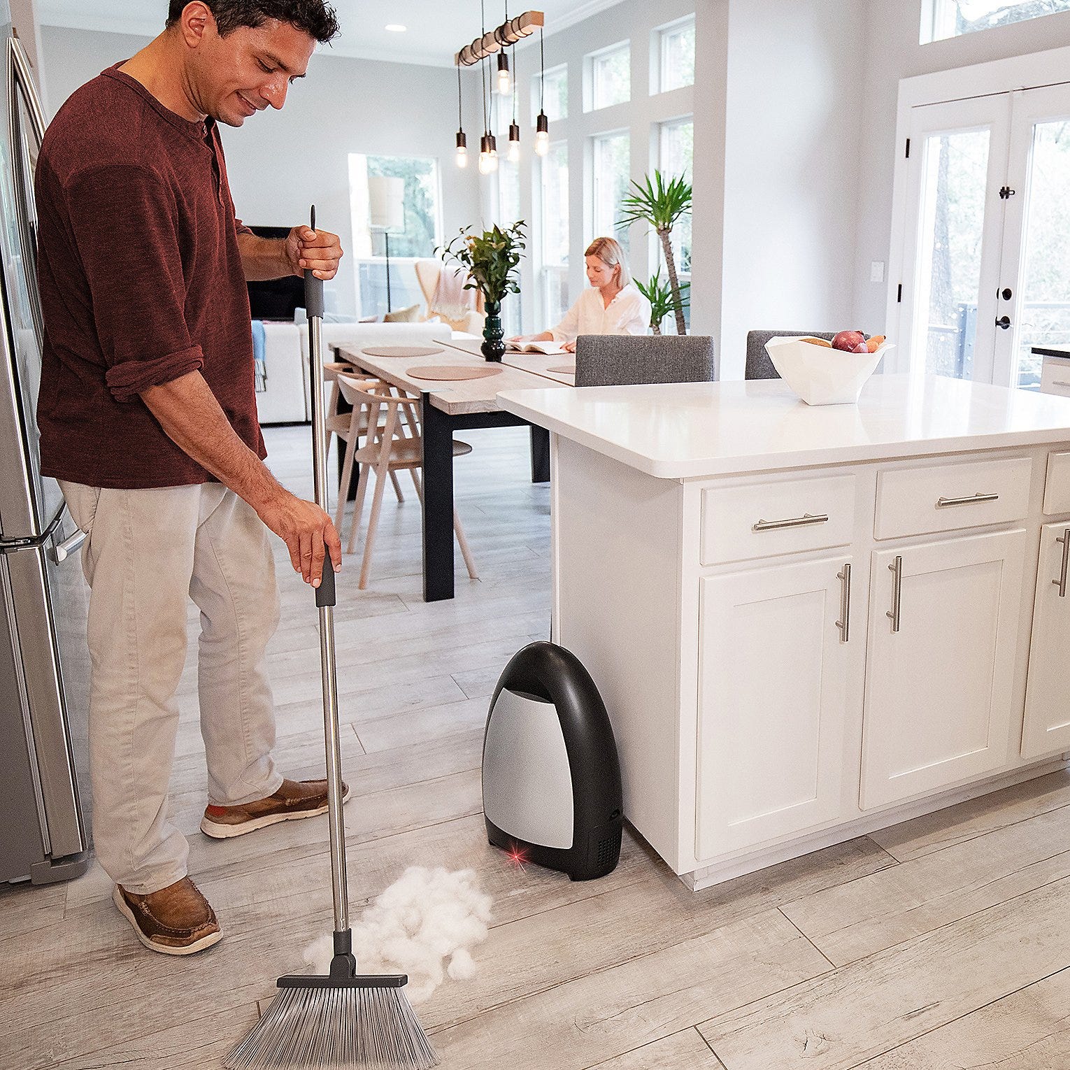 A man sweeps debris into a stationary vacuum device in a kitchen while a woman works at a table in the background.