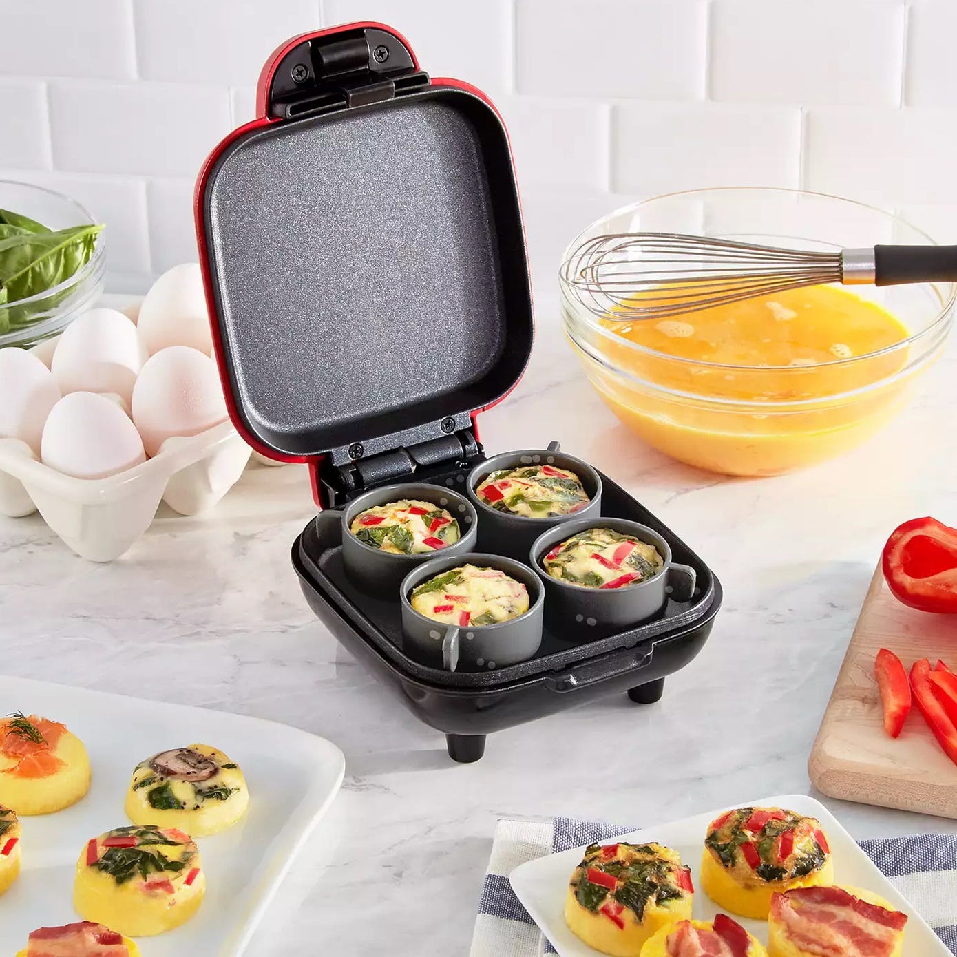 An open egg bite maker with six compartments containing uncooked egg mixture, next to ingredients like eggs, vegetables, and a bowl of whisked eggs.