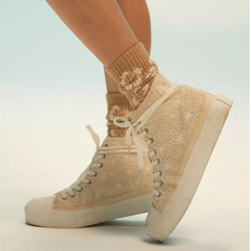 A lace-up, high-top sneaker with a textured, floral pattern in a light beige color, paired with a matching sock.