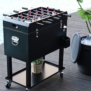 A black, portable foosball table on wheels with extended handles and a lower shelf, placed on a patio.