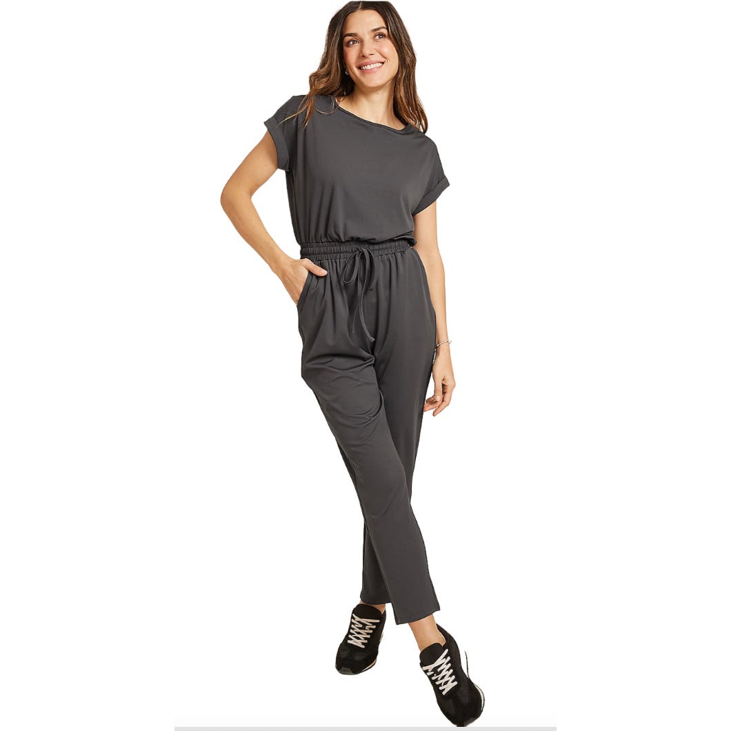 A woman is wearing a dark gray jumpsuit with short sleeves and an elastic waist with a drawstring. The outfit is paired with black sneakers, and the jumpsuit has a relaxed, comfortable fit.