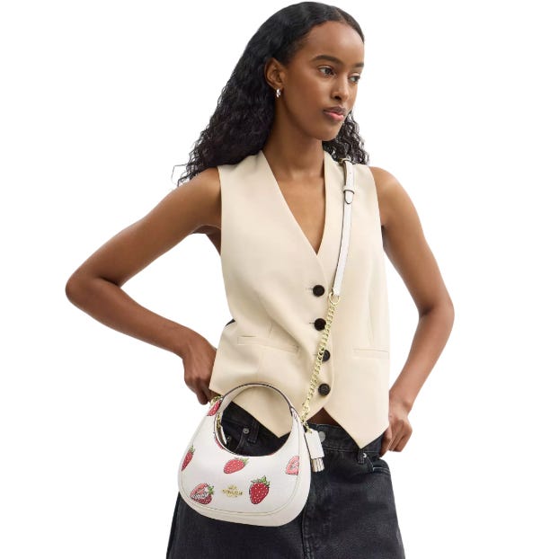 A woman wearing a cream vest and black jeans holds a Coach hobo bag featuring a strawberry print, decorated with a gold-tone chain strap.