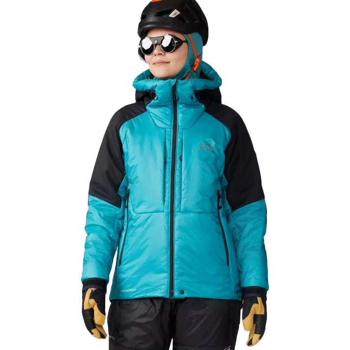 A teal and black Alpine ski jacket featuring a high collar, insulated design, and the Mountain Hardwear logo on the chest. The person is also wearing a helmet, goggles, and gloves.