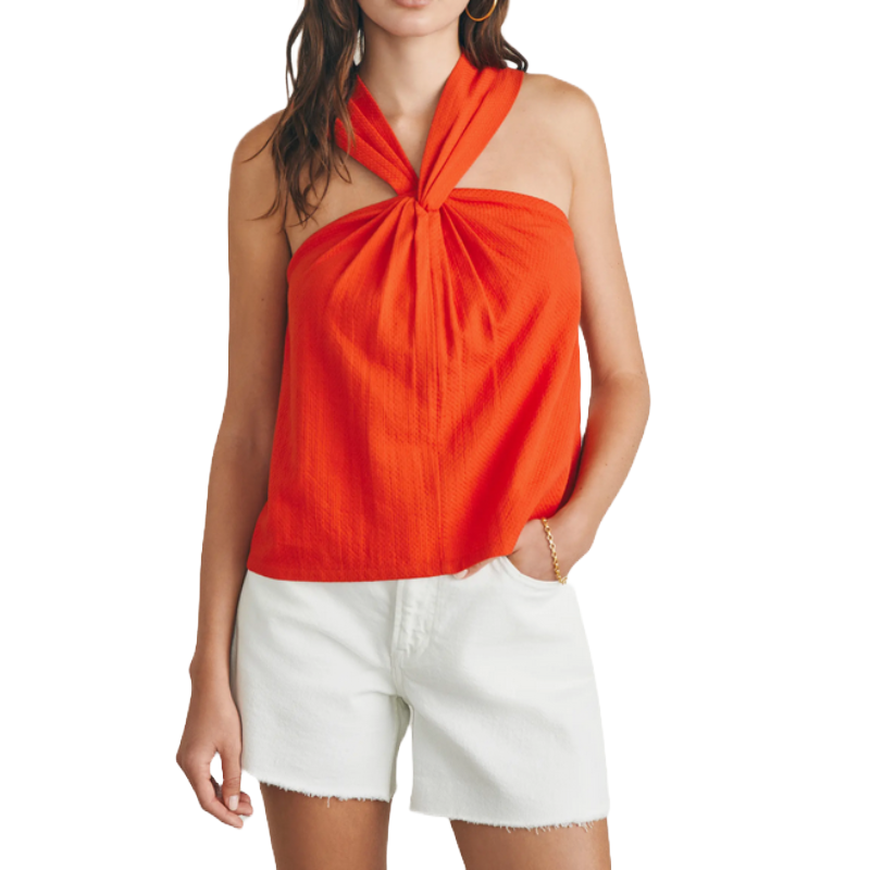 A woman in a vibrant orange halter top paired with white denim shorts stands with her left hand in her pocket, showcasing a casual summer style.