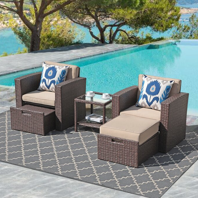 Two wicker chairs with beige cushions, a matching ottoman, and a small glass-topped table on an outdoor rug by a pool.
