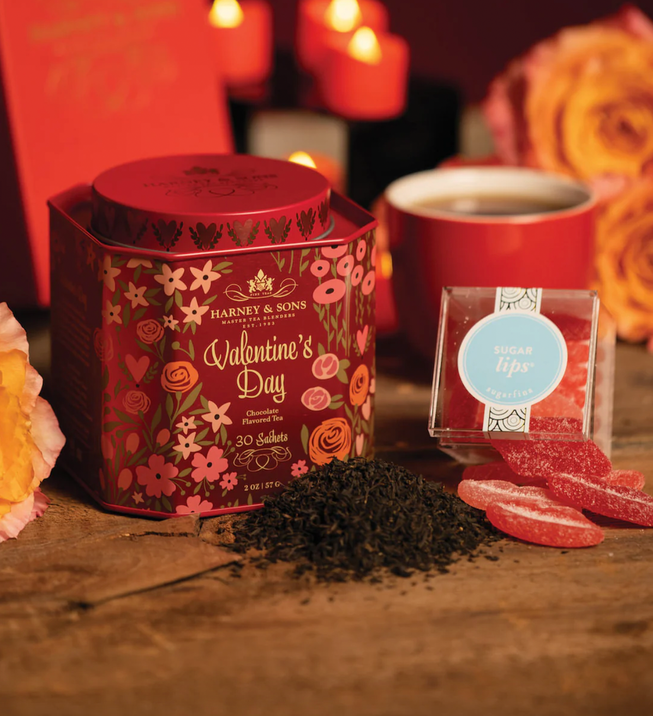 Harney & Sons Valentine's Day Tea Set features a decorative tin of chocolate-flavored tea sachets, displayed alongside a pile of loose tea, a red cup, and Sugarfina \“Sugar Lips\“ gummy candies, all set against a romantic background with candles and roses.