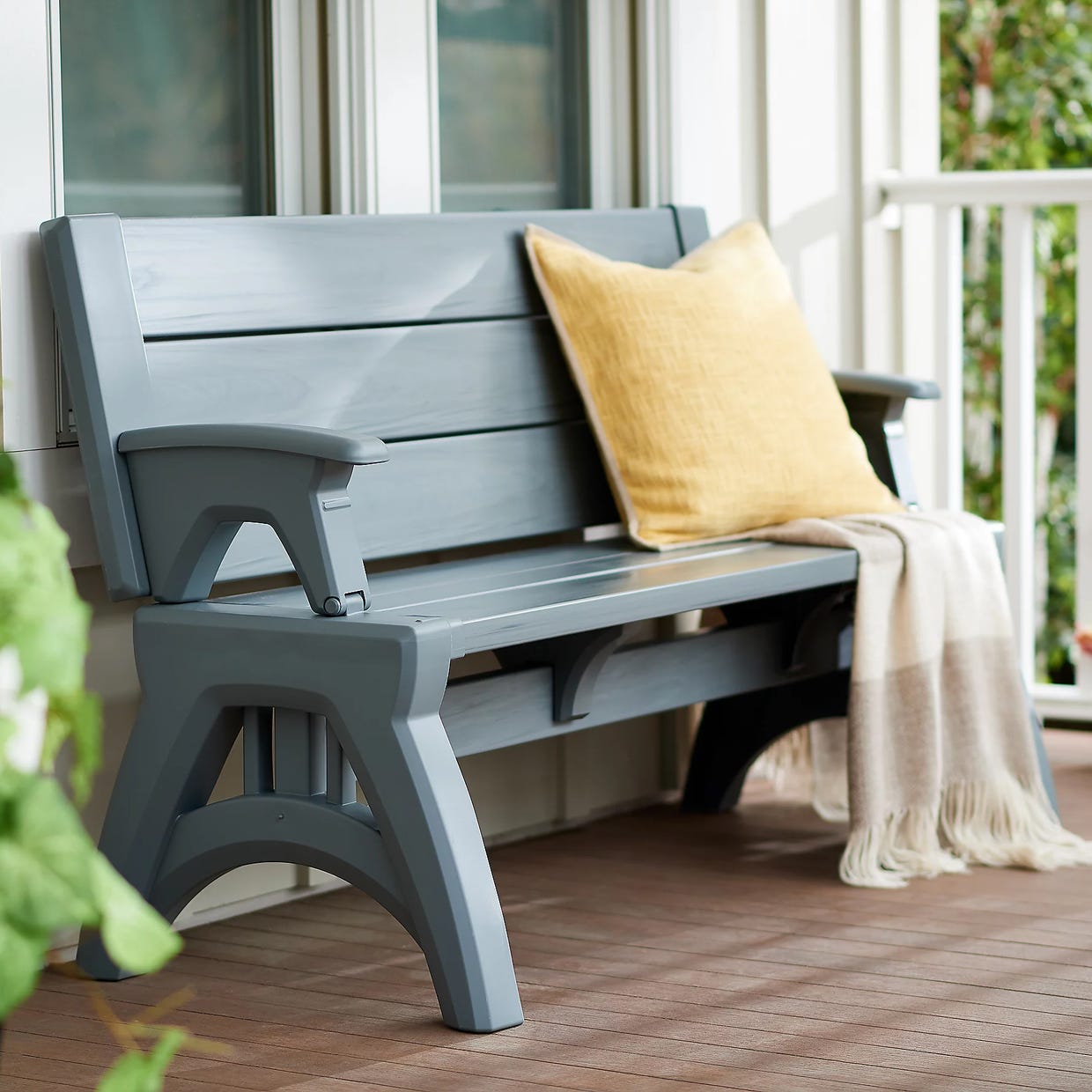 A gray outdoor bench with armrests, a yellow cushion, and a beige throw blanket on a wooden deck.