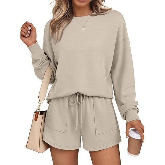 A woman is wearing a beige long-sleeve lounge set consisting of a sweatshirt and drawstring shorts, holding a coffee cup and a handbag.