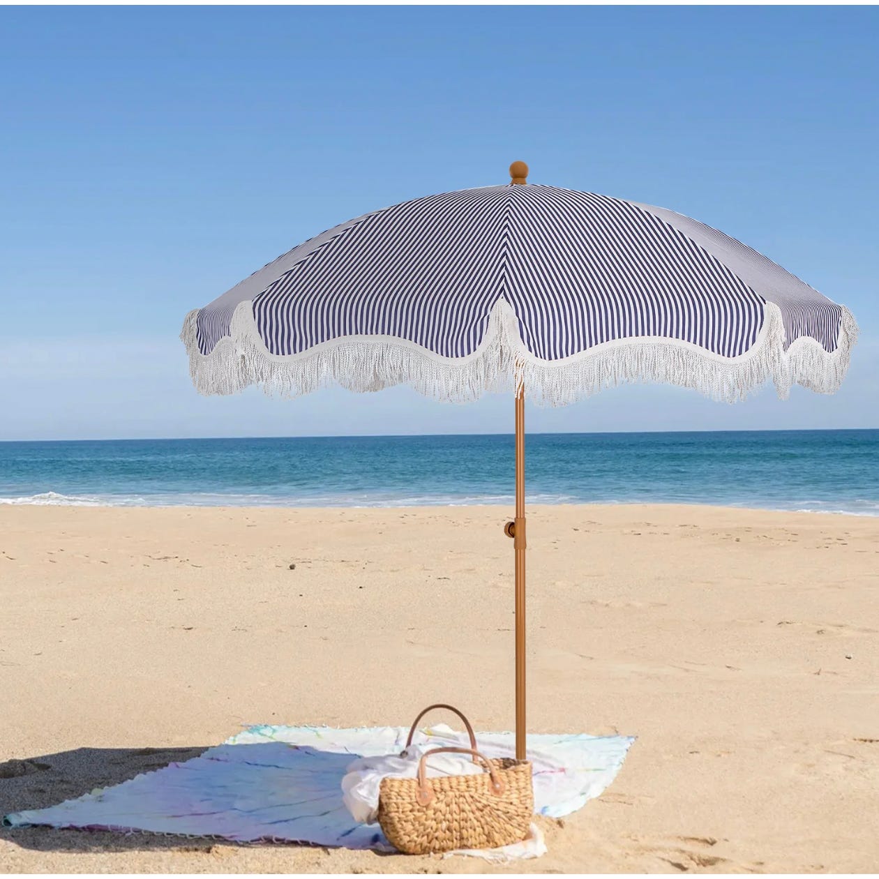 A 7-foot auto-tilt beach umbrella with blue and white stripes and fringe, positioned on a sandy beach with a woven basket and blanket underneath.
