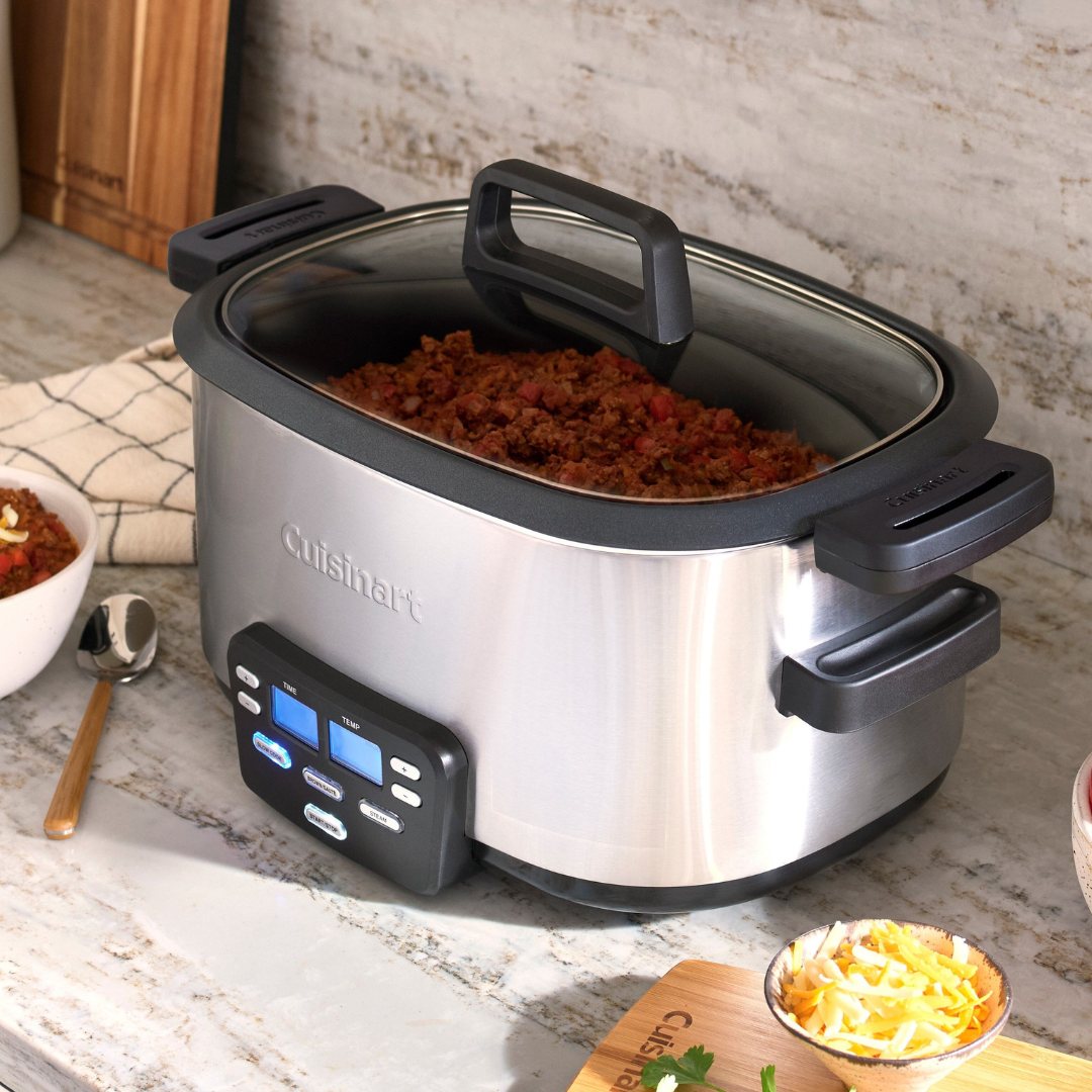 The Cuisinart 6-Qt. Multicooker is a stainless steel kitchen appliance with a digital display and control panel, featuring multiple cooking functions. It's shown here making a dish with cooked meat, alongside bowls and a wooden cutting board with toppings.