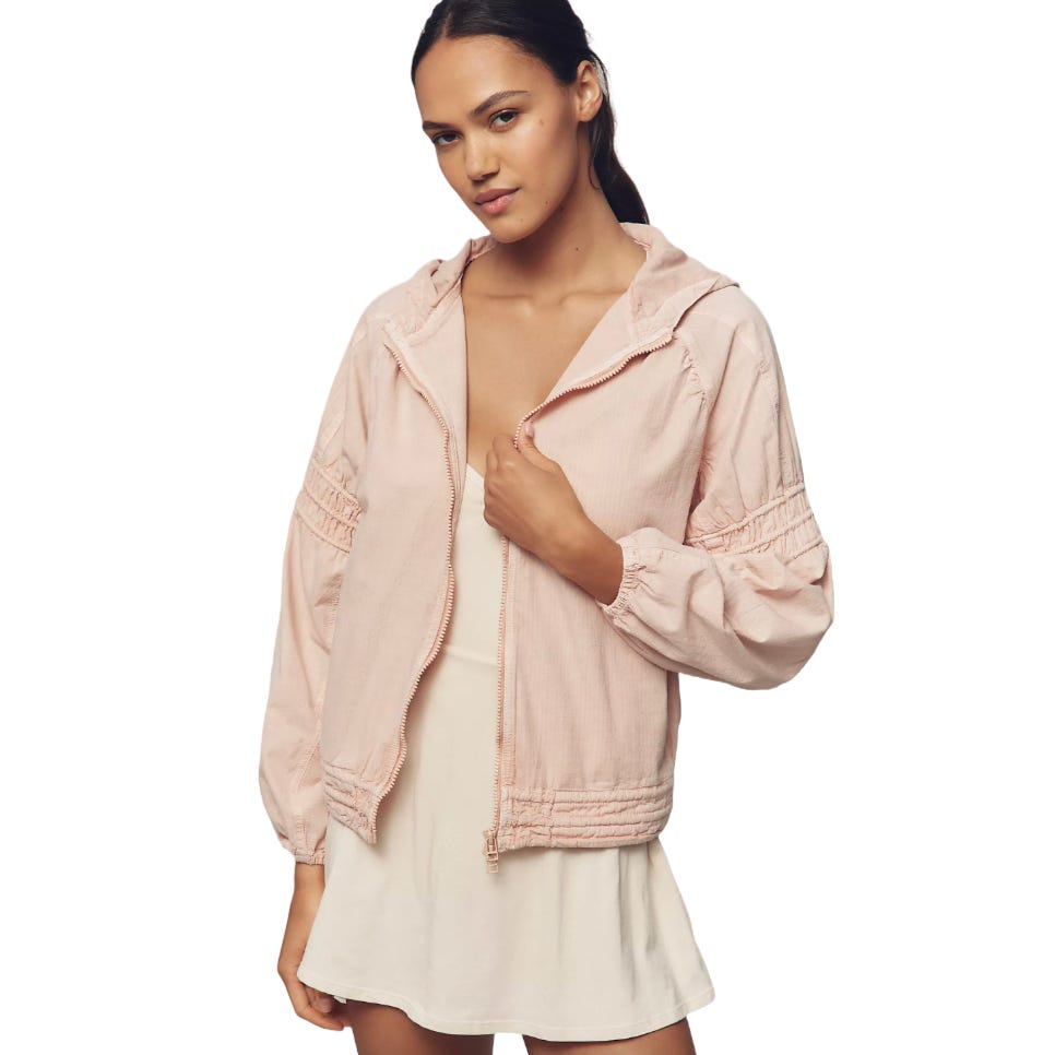 A woman is wearing a light pink, hooded, zip-up jacket over a short, white dress. The jacket features gathered detailing on the sleeves and hem.