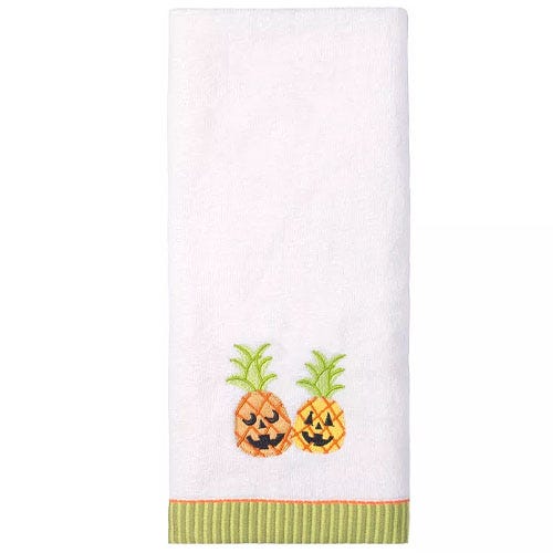 A white kitchen towel featuring embroidered jack-o'-lantern pineapples with greenery on top and a green striped hem accent.