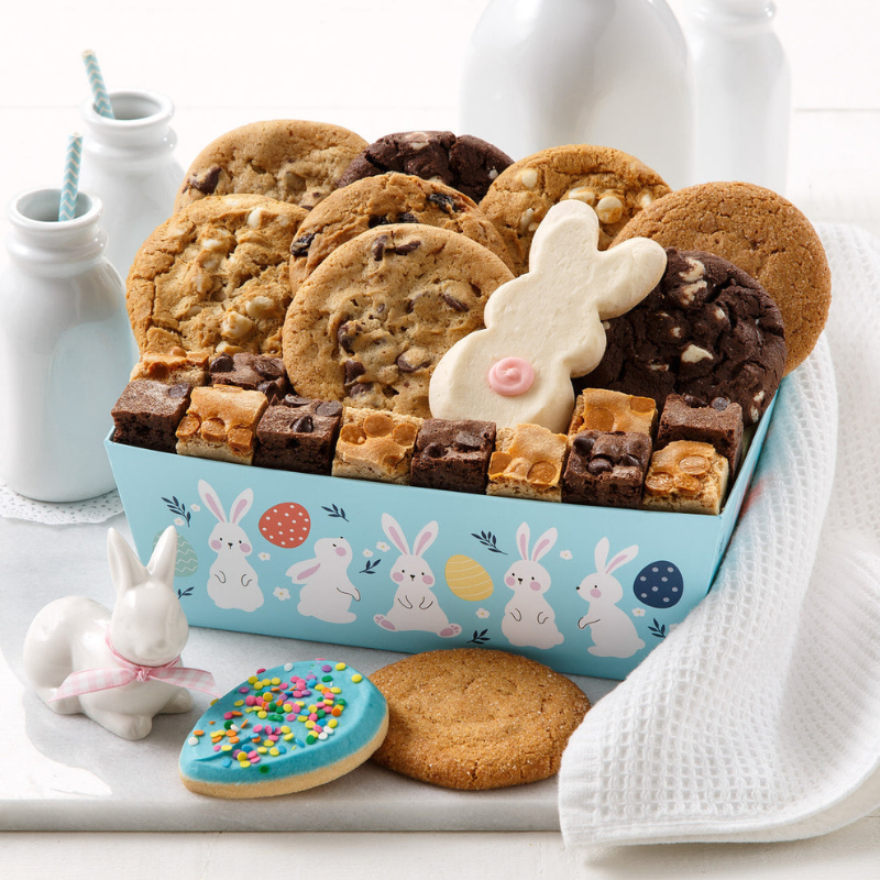 A blue crate decorated with bunnies and eggs contains an assortment of cookies, including chocolate chip, white chocolate, and a bunny-shaped iced cookie, along with mini brownies and blondies. A ceramic bunny and an egg-shaped iced cookie are also shown nearby.