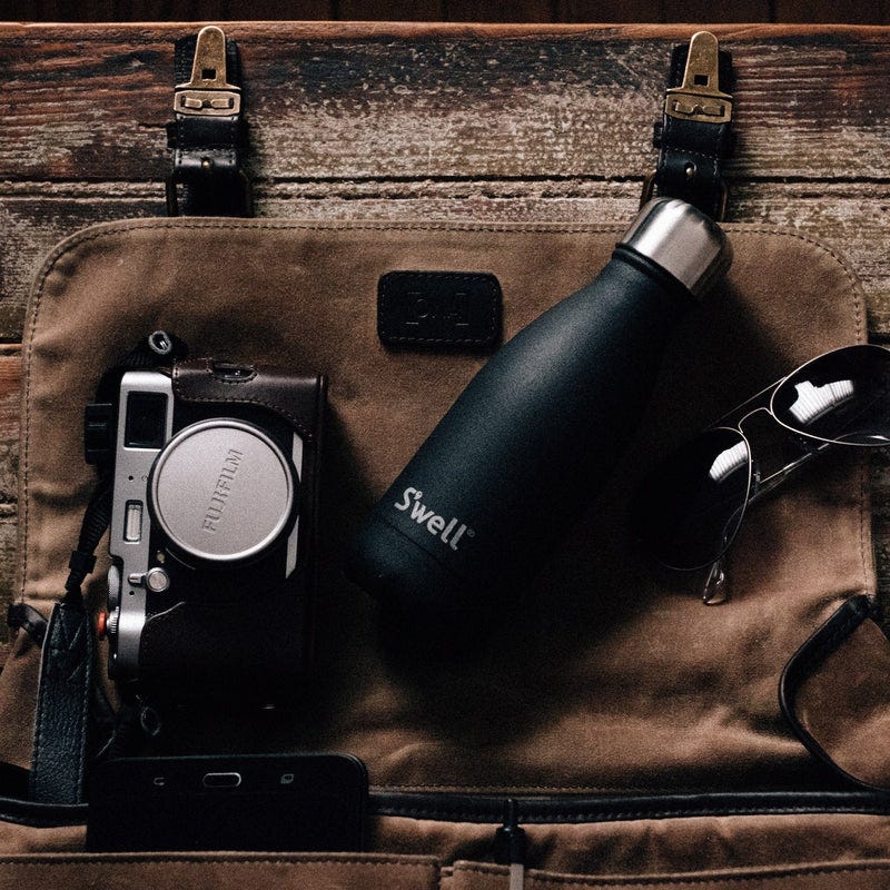 A black 9oz S'well bottle with a matte finish and a silver cap rests beside a camera and earphones on a brown bag.