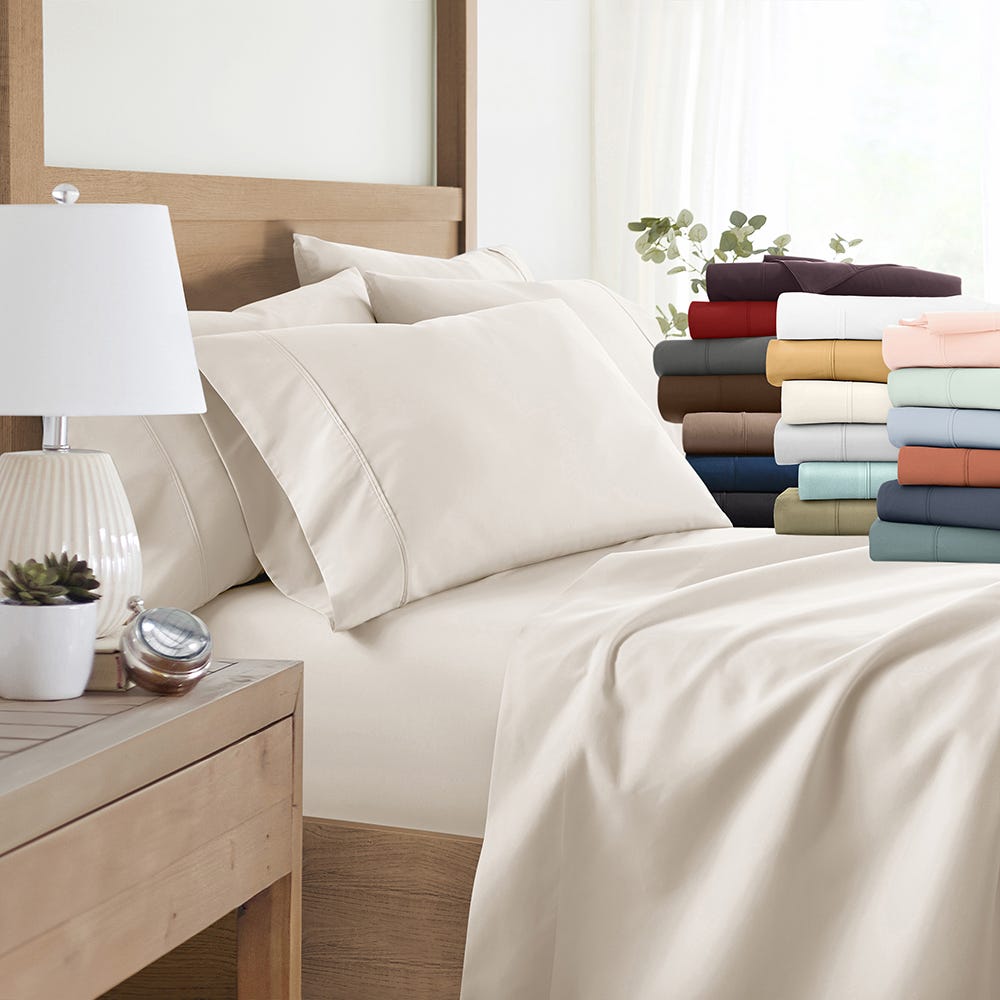 Bed with beige bedding set in a room and an assortment of colored bedsheet sets stacked on the side.