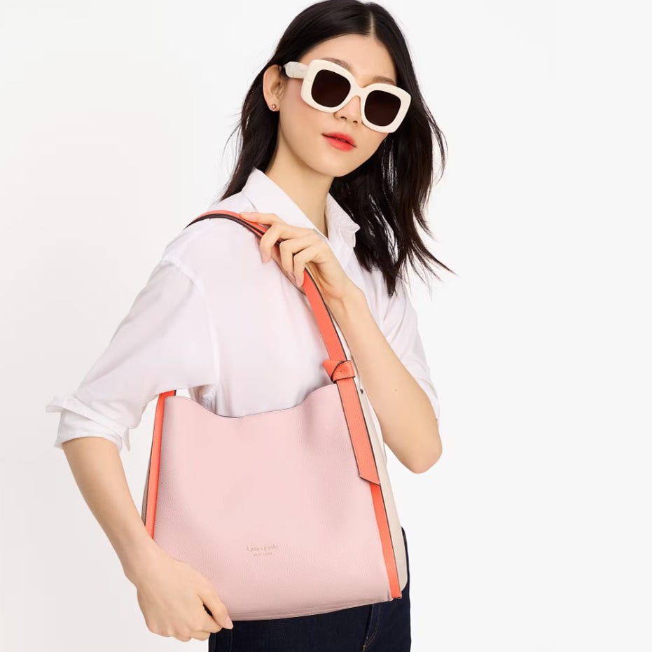 A woman wears white square sunglasses and carries a pink shoulder bag with orange details.