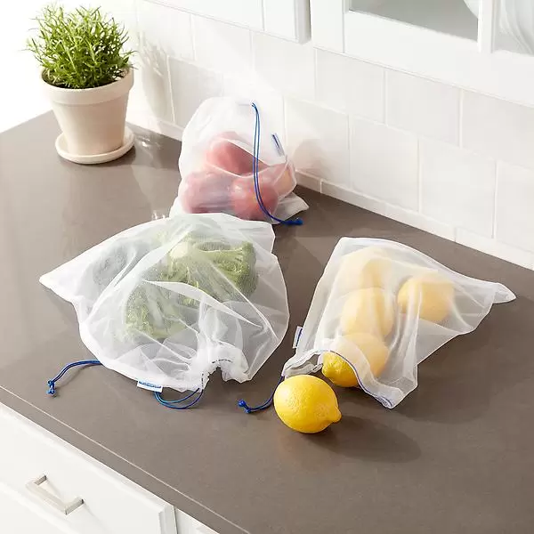 Mesh reusable produce bags with drawstring closures, containing broccoli, lemons, and red peppers. They're placed on a kitchen countertop next to a potted plant.