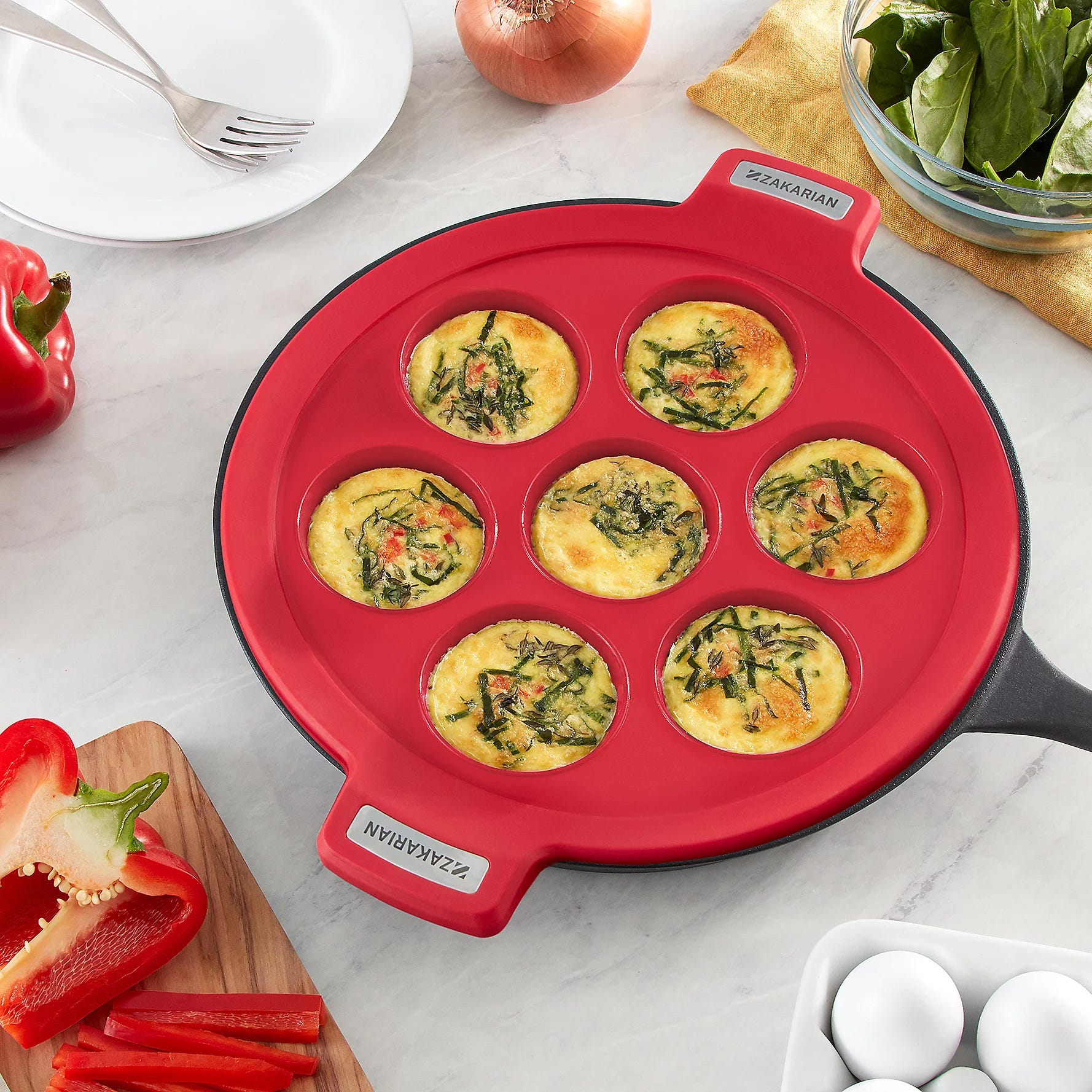 Red silicone egg bite mold with multiple compartments filled with cooked spinach and red pepper egg bites, used on a stovetop.