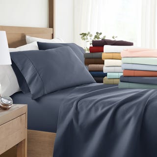 Assorted bed sheets and pillowcases in various colors neatly stacked with the bed partially visible.