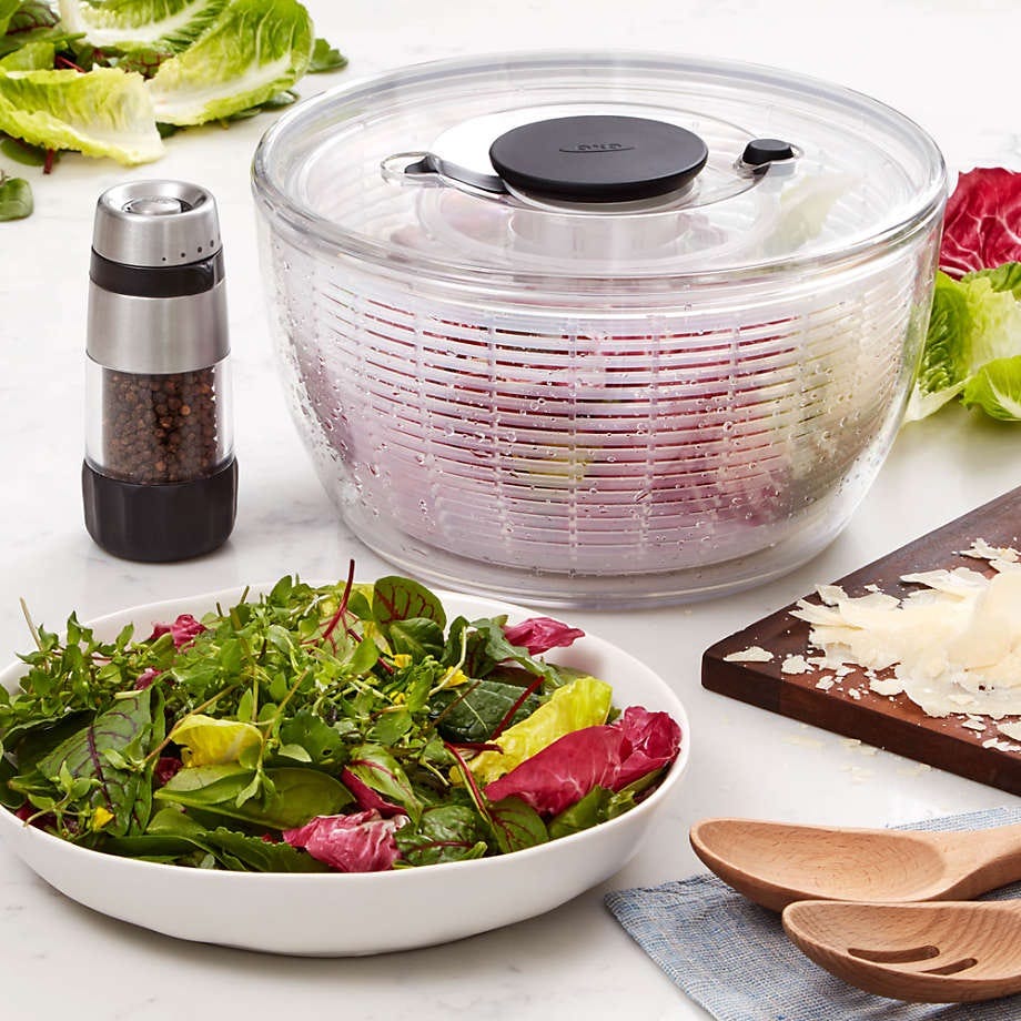 A salad spinner, mixed greens in a bowl, a pepper grinder, cheese on a cutting board, and wooden salad utensils.