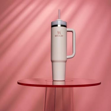 A 32oz Stanley tumbler in a muted color with a straw, a handle, and the Stanley logo on its side, set against a rose-colored backdrop with diagonal lines.