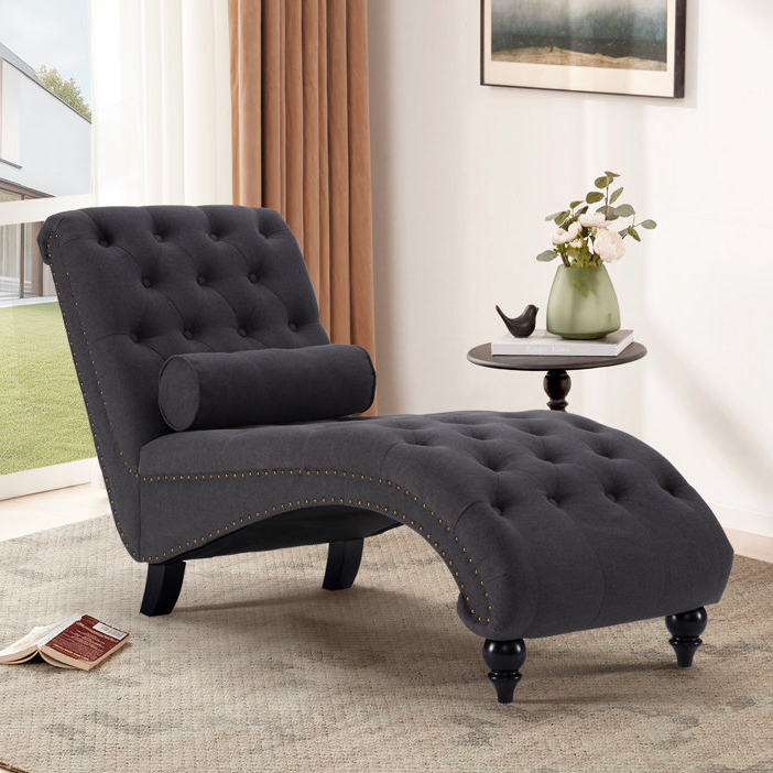 A dark gray tufted chaise lounge with a matching accent pillow and studded detailing, next to a small round side table with a vase of flowers.