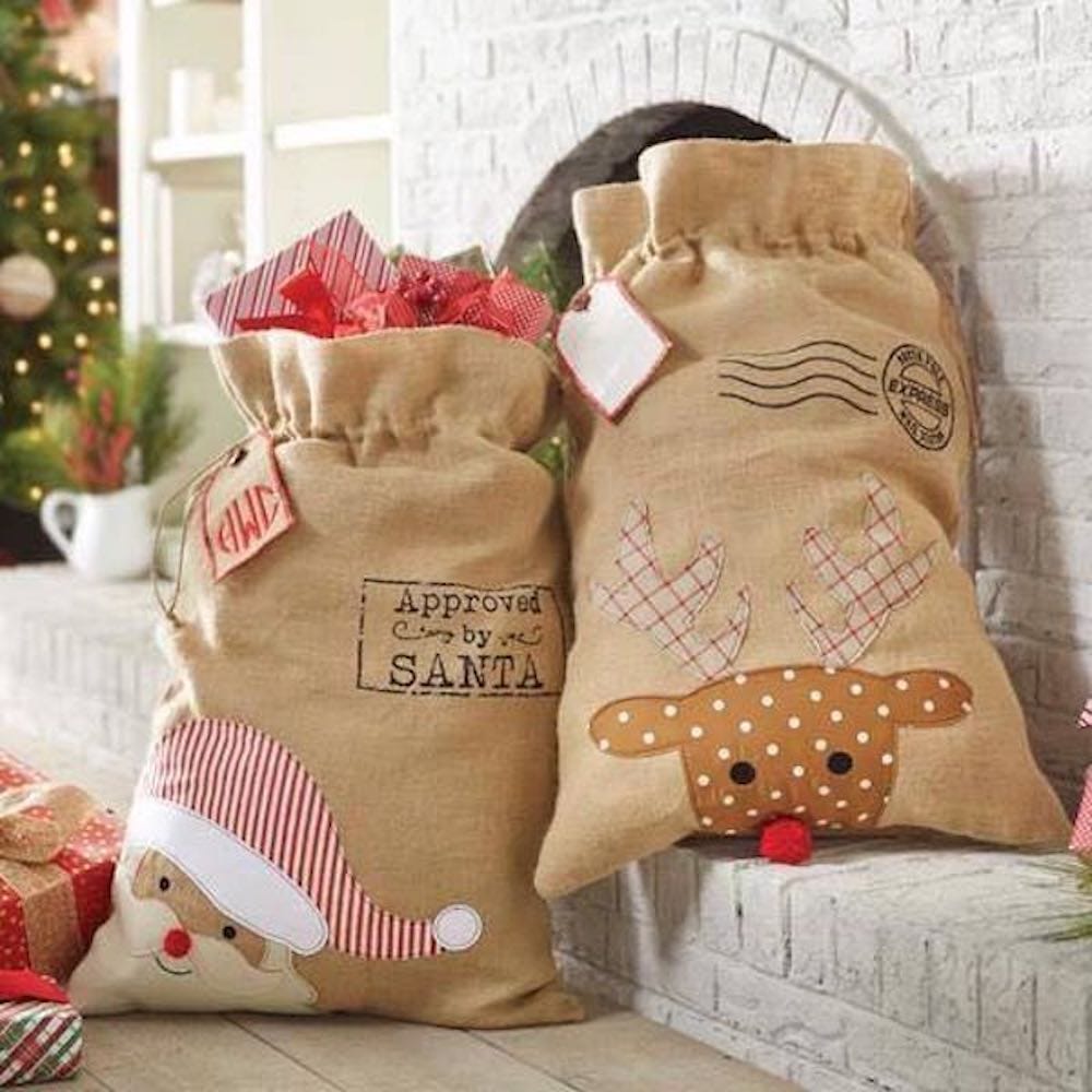 Two burlap drawstring sacks sit by a fireplace, one featuring a Santa Claus design labeled \“Approved by Santa,\“ and the other decorated with a reindeer face with plaid antlers and a red nose. Both sacks are filled with gifts and have festive tags attached.
