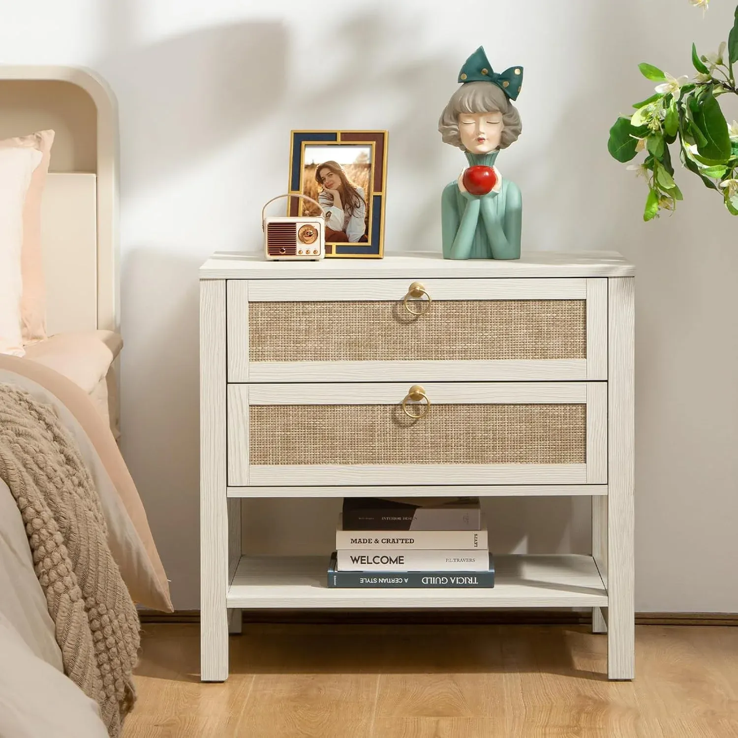 The Terra White Nightstand features a minimalist design with two drawers and woven rattan fronts, adorned with round metallic handles. A lower shelf holds stacked books, and the top surface displays a small vintage-style radio, a picture frame, and a figurine.