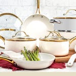 A set of cream-colored cookware with gold handles, including pots, a pan, and utensils, displayed on a kitchen counter.