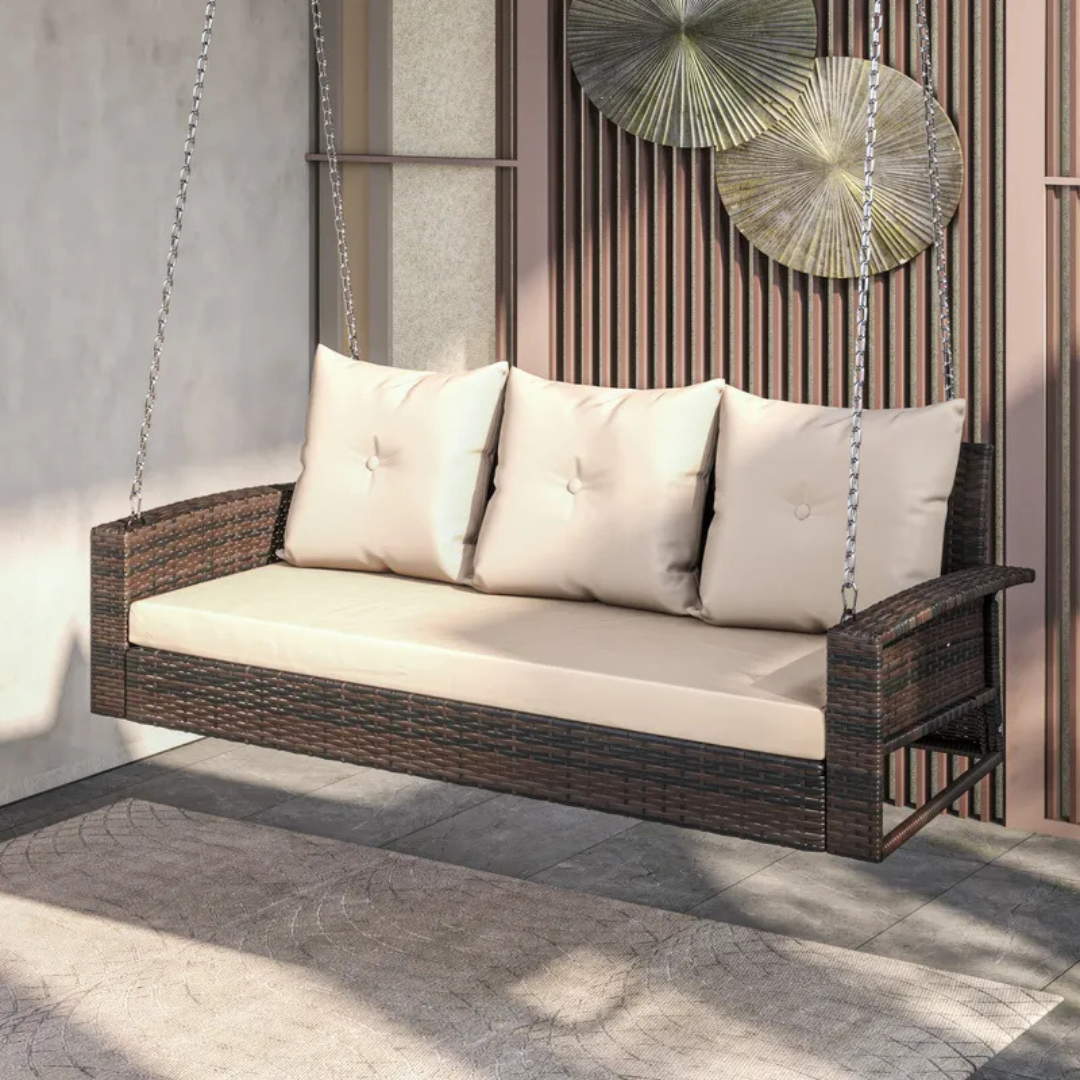 A 3-person porch swing with a dark brown wicker frame, beige cushions, and back pillows, suspended by metal chains. The swing features an armrest on each side and is positioned against a wall with decorative elements.