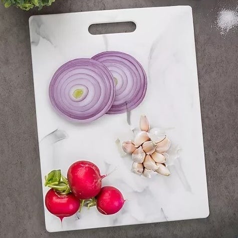 A white marble-patterned cutting board with a handle displays sliced red onions, peeled garlic cloves, and radishes, set against a dark surface.