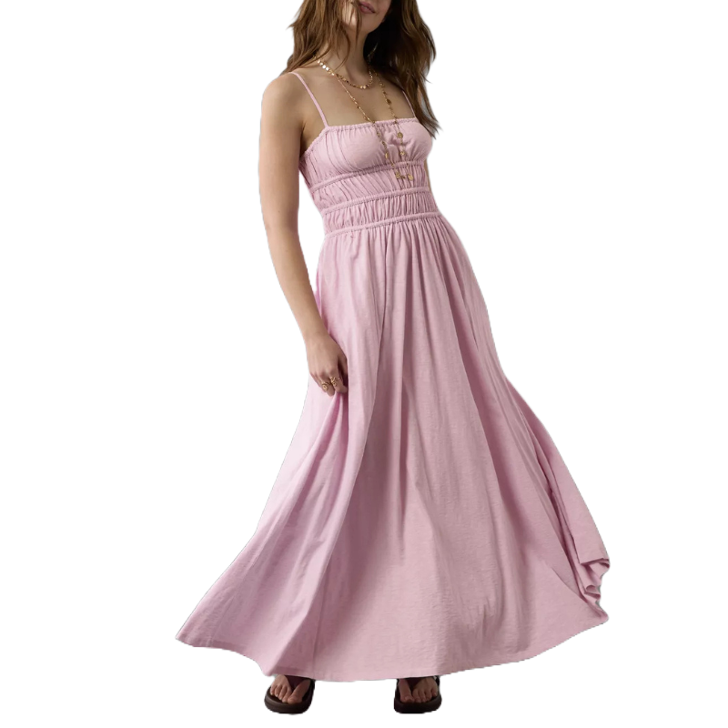 A woman is wearing a light pink maxi dress with spaghetti straps and a gathered bodice, paired with dark sandals.