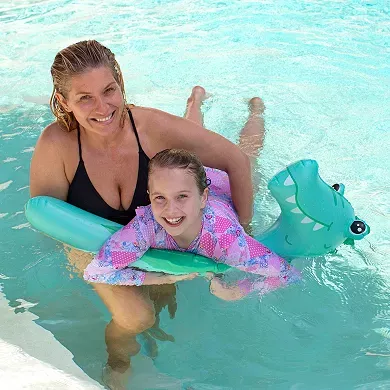 A smiling adult and child are in a pool; the child is supported by a green frog-shaped pool noodle and wearing a pink-patterned flotation vest.
