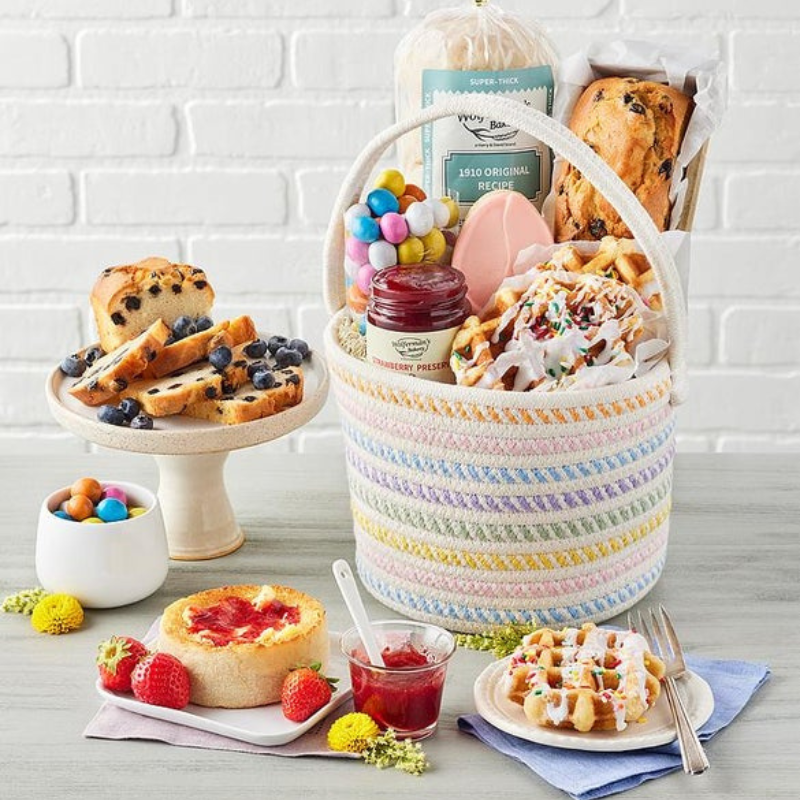 A colorful basket contains a loaf of bread, a jar of strawberry preserves, a blueberry loaf, waffles with icing and sprinkles, pastel egg candies, and a decorated cookie. Nearby are a dessert plate with jam and strawberries, and a stand with sliced blueberry loaf.