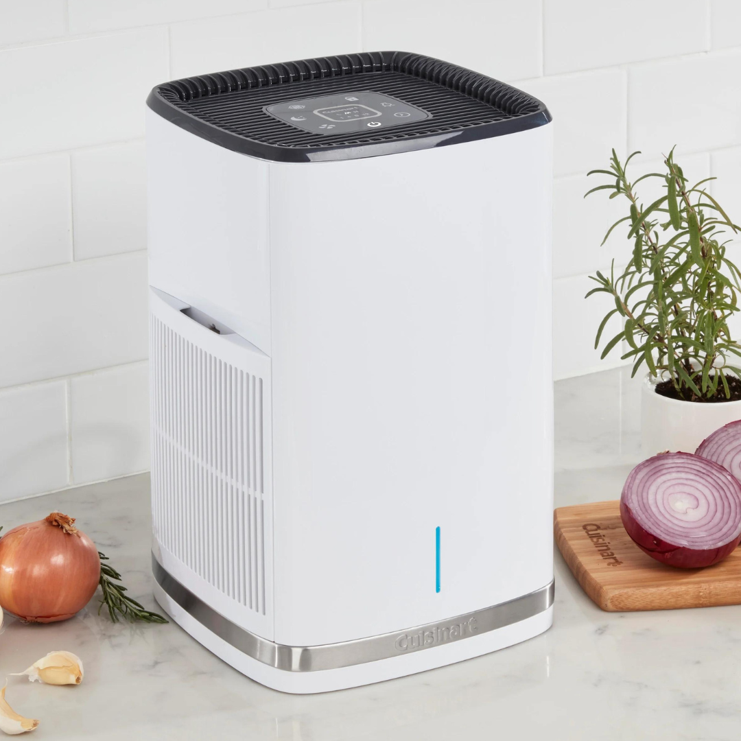 A white air purifier with a digital control panel on top, sitting on a kitchen counter next to herbs and sliced onions.