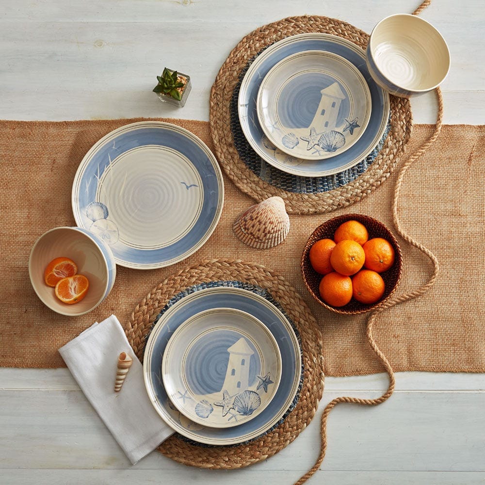 The 12pc Rio Beach House Melamine Dinnerware features plates and bowls with coastal designs, including seashells and a lighthouse in muted blues and creams. They are displayed on woven placemats with a beach-themed décor, alongside oranges and a succulent.