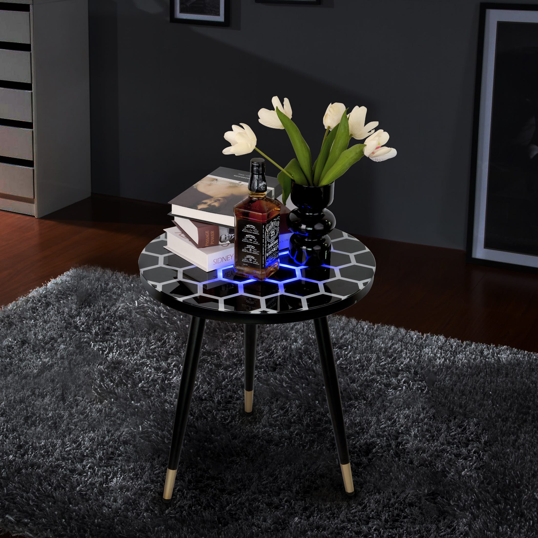 A round side table with a hexagonal patterned top, illuminated with blue LED lights, displaying books, a black vase with white tulips, and a cologne bottle.
