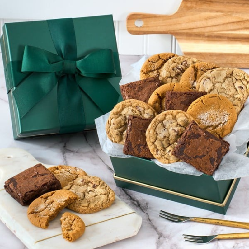 A green gift box with a ribbon contains an assortment of cookies and brownies, including chocolate chip and sugar cookies accompanied by dense chocolate brownies. A few treats are displayed on a marble board nearby.