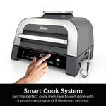 A Ninja Foodi XL Cooker with a digital display and a person's hand touching the control panel. Features include a Smart Cook System with 4 protein and 9 doneness settings.