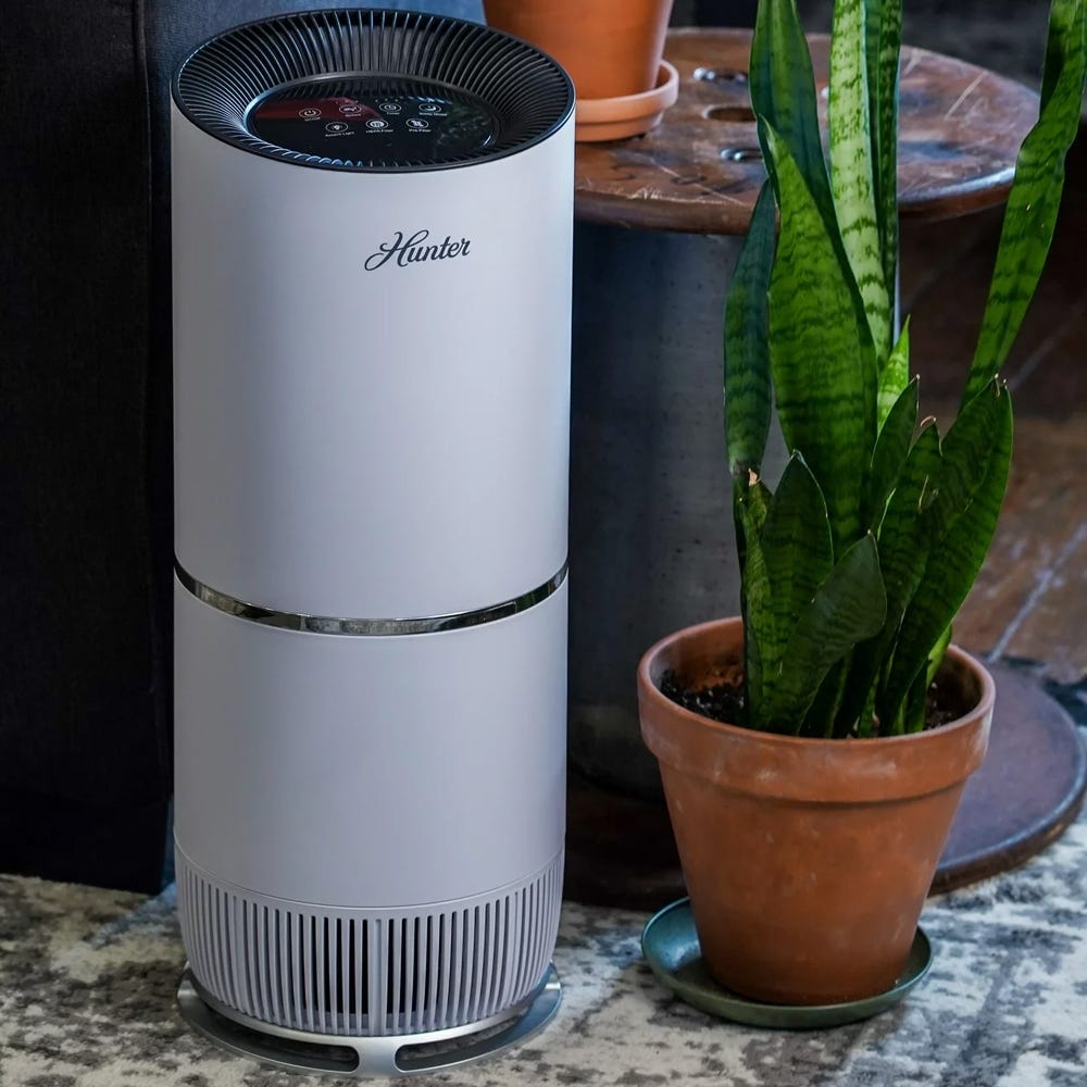 A white air purifier with a digital control panel on top, next to a potted snake plant.