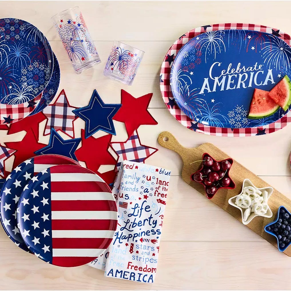 Patriotic-themed partyware including plates, cups, and star-shaped decorations in red, white, and blue. There are also fruit snacks arranged on cutting boards.