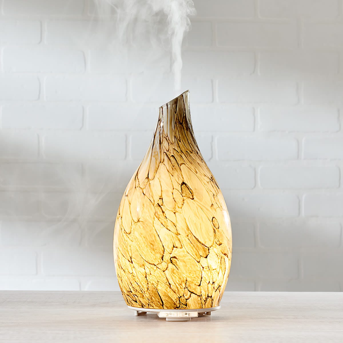 A sand-blasted glass diffuser with a marbled pattern emits mist from a narrow, angled top, creating an illuminated, ambient glow. It's elegantly rounded and sits on a flat base.