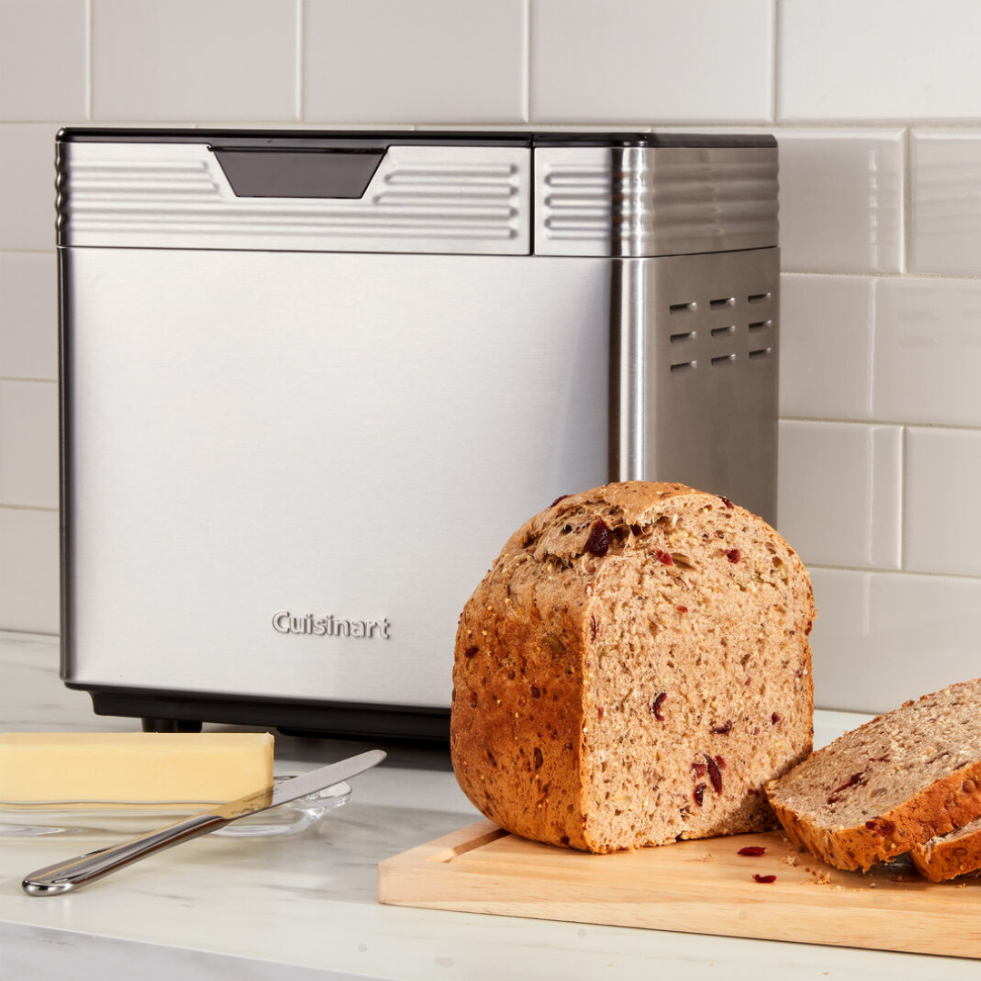 Cuisinart Bread Maker with a sleek stainless steel exterior is shown next to a loaf of multigrain bread and a stick of butter. It's positioned on a kitchen countertop with tiled backsplash.