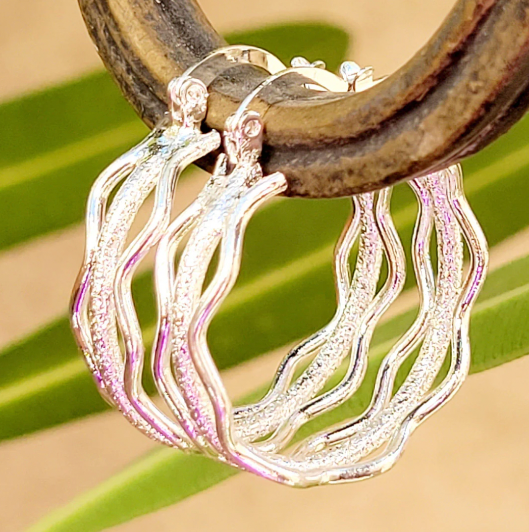 Silver wavy hoops with a textured design, featuring three intertwined waves. They have a shiny finish and are hanging from a wooden display.