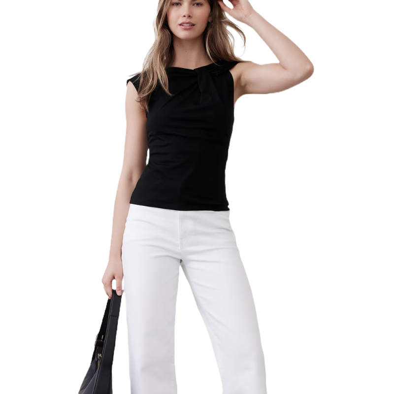 A woman wearing a sleeveless black top with a bow detail, paired with white pants. She is holding a black bag.