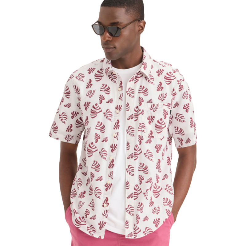 A man wearing a short-sleeve button-up shirt featuring a pattern of red-striped seashells on a white background, paired with a white T-shirt and pink shorts. He also has on dark sunglasses.