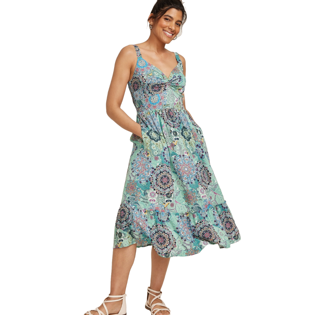 A woman is wearing a sleeveless, knee-length dress with a colorful floral and paisley pattern, paired with strappy sandals.