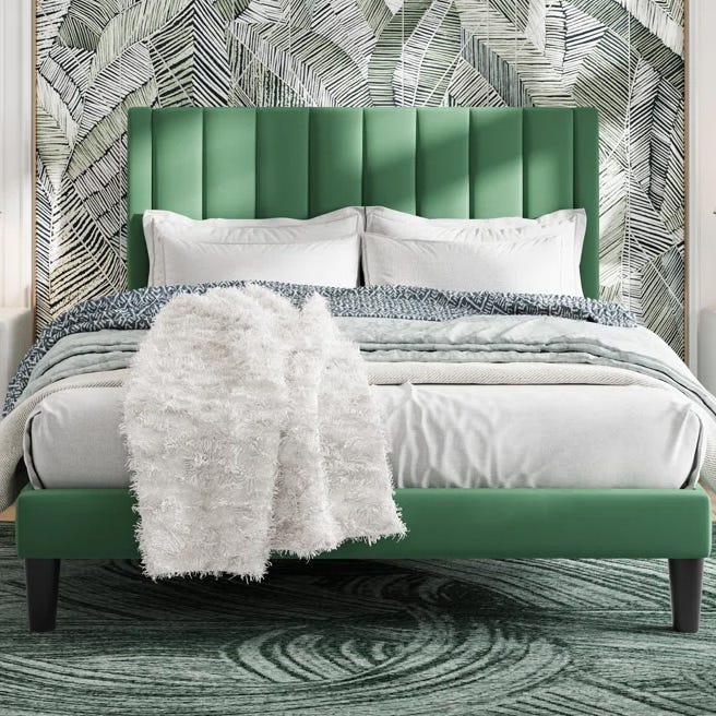 A bed with a green upholstered headboard is set against a patterned leaf wallpaper. It features white bedding, a textured white throw, and a green circular pattern rug.