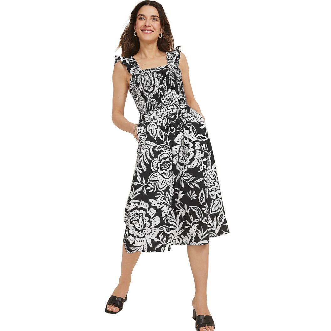 A woman is wearing a sleeveless black dress with a white floral pattern, featuring ruffled straps and a midi length. She completes the look with black open-toe sandals.