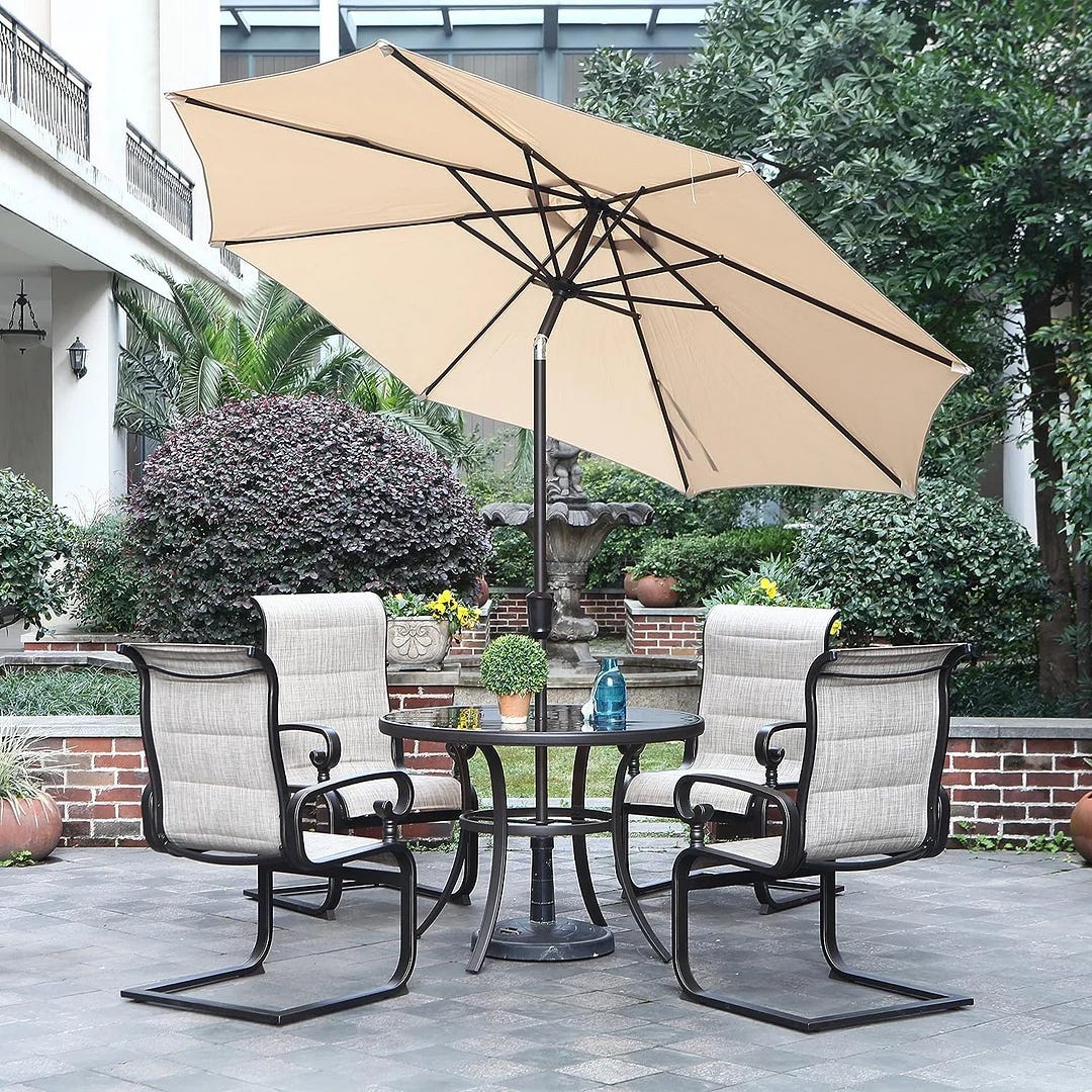 A patio set with four chairs and a round table under a beige umbrella, arranged on a paved outdoor area.